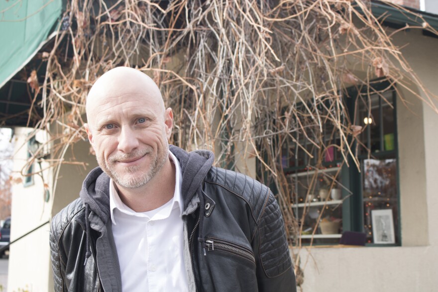 A man in a black leather jacket grins, as he gently leans to one side while standing in front of a tree. 