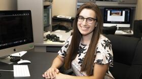 A woman with brown hair, and glasses, sitting down, smiling at the camera.