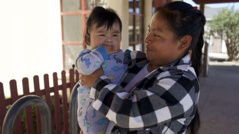 A woman holds her baby, who is wearing a blue onesie with blue and yellow animals.