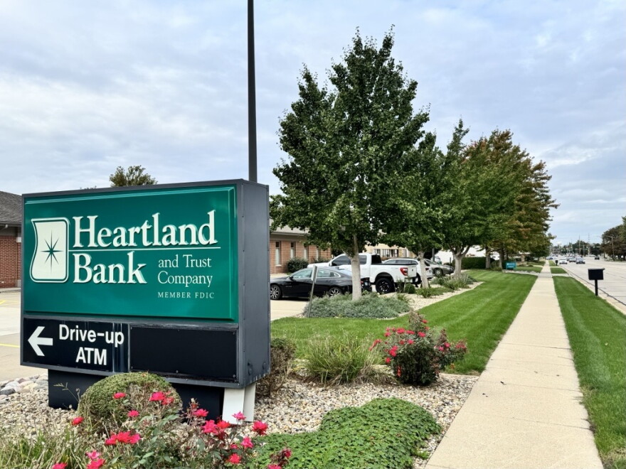 A Heartland Bank and Trust sign directs customers toward the ATM in front of a row of trees.