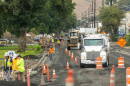 A large white truck surrounded by orange traffic cones and men in safety vests and hard hats in the foreground show construction underway on Snake River Avenue in Lewiston. 