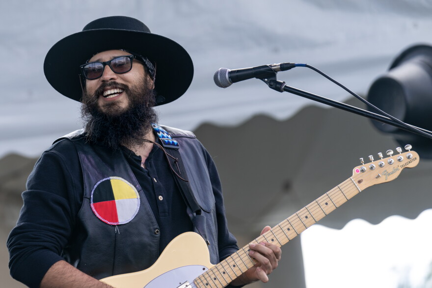 A smiling man playing guitar on stage. He is wearing a wide brimmed black hat, a black shirt and a black vest with a black, yellow, red and white patch on it.