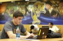 A sophomore at Florida International University types on his laptop in a common hall with the FIU Panther logo behind him.