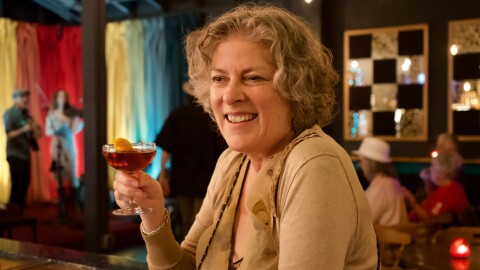 A woman holds a cocktail and smiles leaning on a bar with a busy scene behind her. 