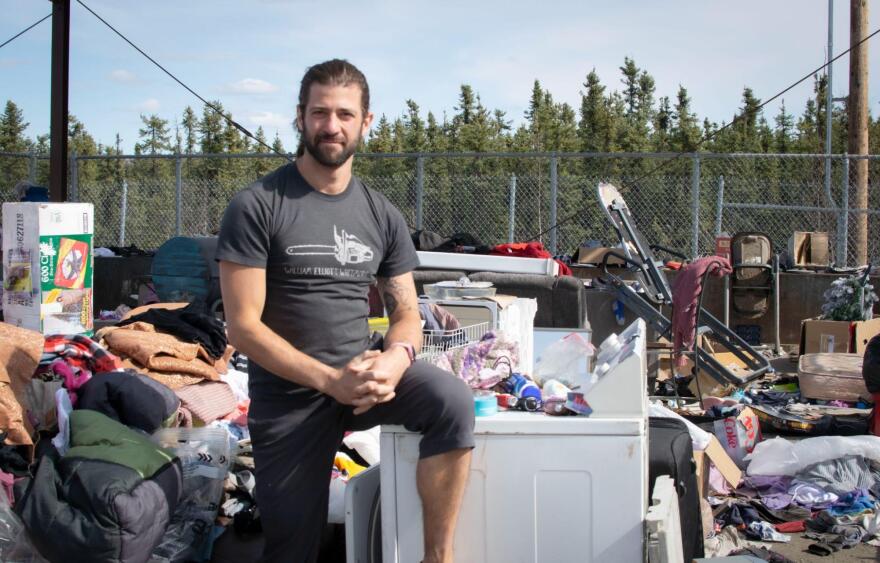 A man wearing a t-shirt with a chainsaw on it stands with one leg propped on a discarded washing machine, at a dump.