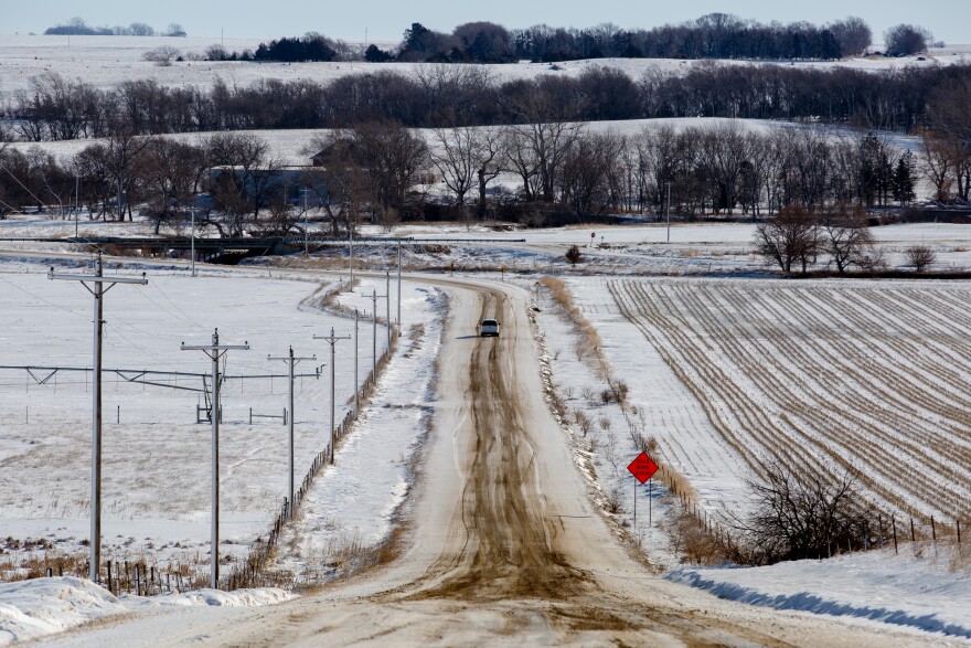 A white van drives down a snowy gravel road, farm fields and pastures surround it.