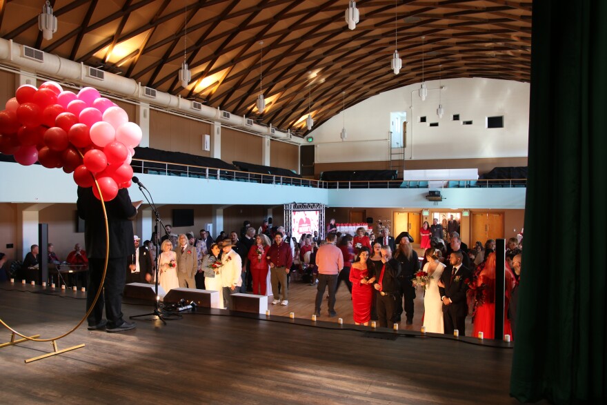 A man is standing in front of a large circular altar with pink and red balloons on it on a stage. He is speaking into a mic. Below him on the ground are several couples, dressed to the nines, listening to him. Behind them are people in the audience in chairs. 