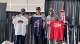 Four men standing on a stage holding up t-shirts with a grey curtain behind them.