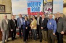 Nearly a dozen men and women stand in front of a sign that reads "Cruise McLean County on the Mother Road"  underneath logos that read "VisitBN" "Enjoy Illinois" 