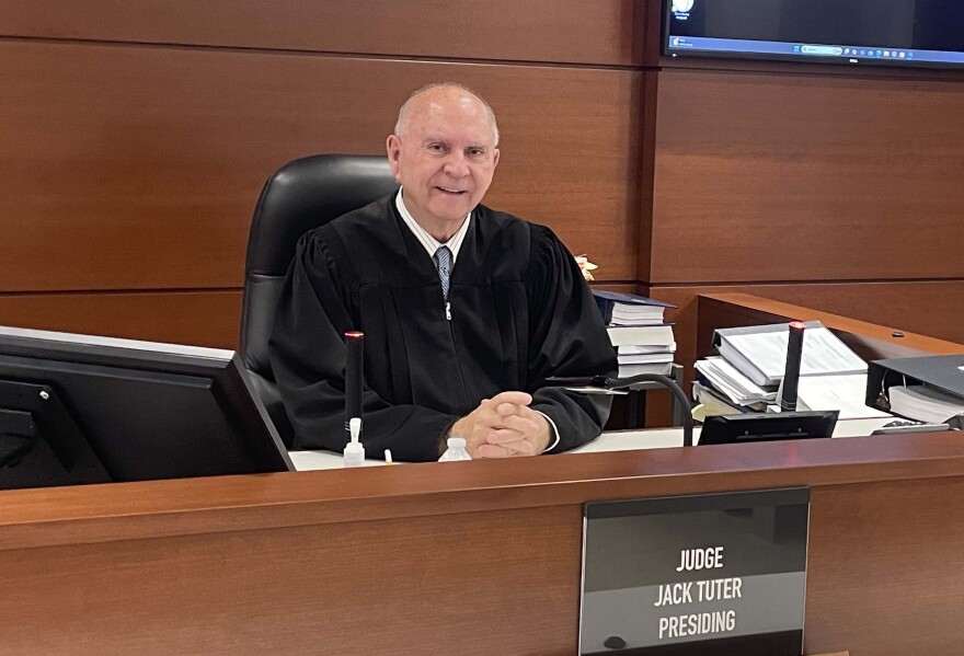 An older man sits in a judge's chair wearing a robe.
