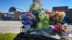 Outdoor photo shows a pile of flowers and stuff animals collecting on top of a concrete pillar. Behind, across the street, is a school building.
