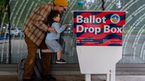 FILE - Matt lifts up his three-year-old son, Theo, to cast his ballot into the Hillsboro Public Library-Shute Park ballot drop box in May 2026. Election officials are increasingly recommending voters plan to use drop boxes rather than mailing their ballot.