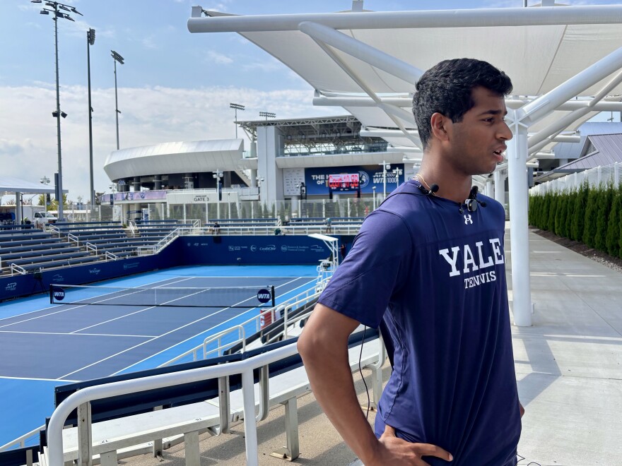 A man in a blue shirt that says "Yale Tennis" in front of an empty blue tennis court and stands.