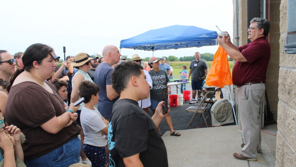 A man stands in front of a crowd holding a weather instrument. He is explaining to the crowd weather balloons work right before he launches one.