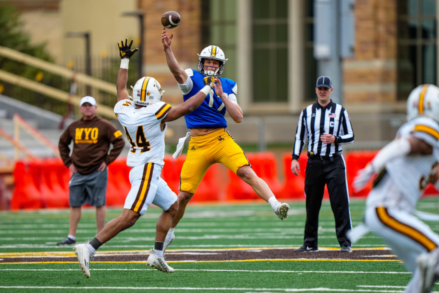 A football payer throws a football while another tries to block it. A coach and a referee look on.