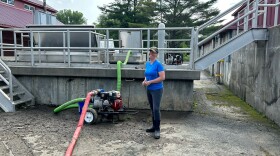 Liz Royer, wearing blue jeans, blue shirt and a camo hat, stands in on mud cover asphalt. There is a water pump machine next to her. 