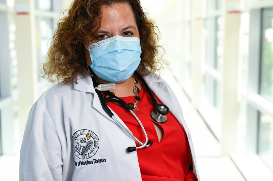 A doctor wearing a mask, white coat and stethoscope stands in a hospital hallway facing the camera.