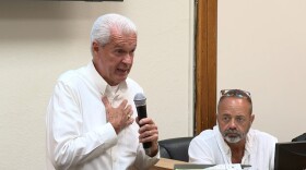 A white haired man speaking into a microphone standing beside a seated bearded man with glasses on his head
