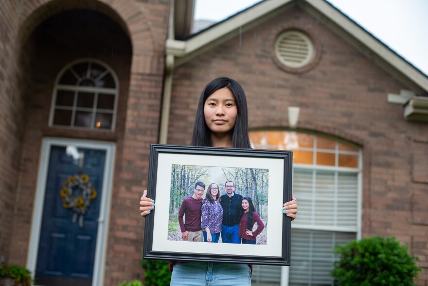 Cosette holds a framed family photo of her adopted parents Tonya Eisenhauer and  John Eisenhauer, and her sibling, Daniel Eisenhauer, in front of their house in Euless, TX., on May 24, 2021.