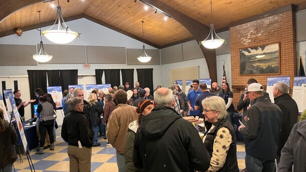38 people of various ages stand in a room talking. Posters and informational tables are set up around the room, and some people working at the tables are explaining things to others with questions.