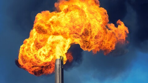 A flare of on-fire gases explodes from a pipe against a blue sky.