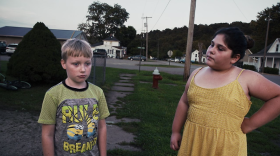 Two children stand on a neighborhood street with a crumbling sidewalk. 