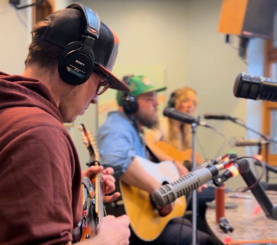 Three musicians looking at their instruments and performing. The nearest is in focus and the other two are slightly blurred.