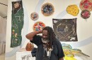 Artist Paul Chandler smiles while standing in front of a display of his mixed-media artwork at the Buffalo AKG Art Museum. The wall behind him is filled with colorful embroidered and beaded textile pieces, alongside sculptures on pedestals. 