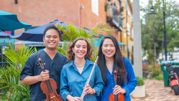 Three young people hold their musical instruments while standing on the sidewalk. 