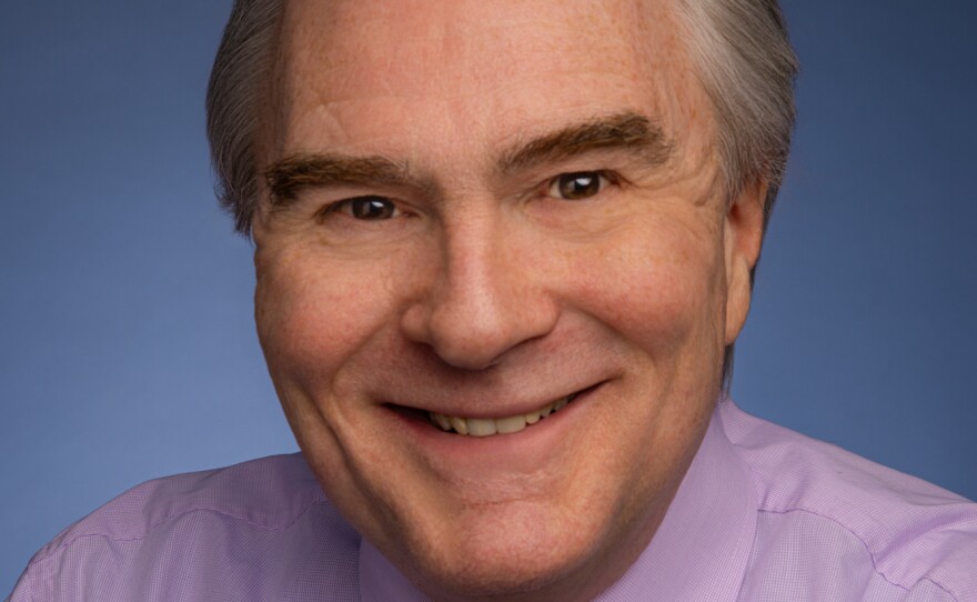 A smiling man with short grey hair is wearing a light purple button-down shirt and dark purple tie.