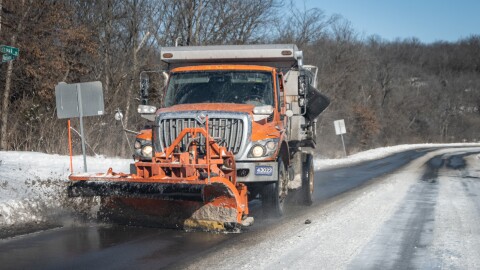 An orange dump truck with a plow attached to the front drives on a partially snow-covered roadway. Slush can be seen flying up from one corner of the plow.