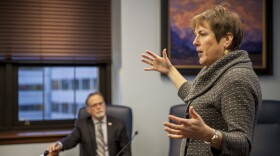 A woman in a grey sweater and short reddish hair gestures in front of a white man at a desk