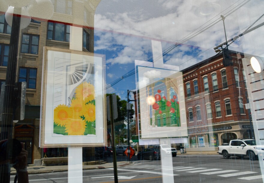 A downtown window reflects the cityscape over two colorful framed prints of flowers.