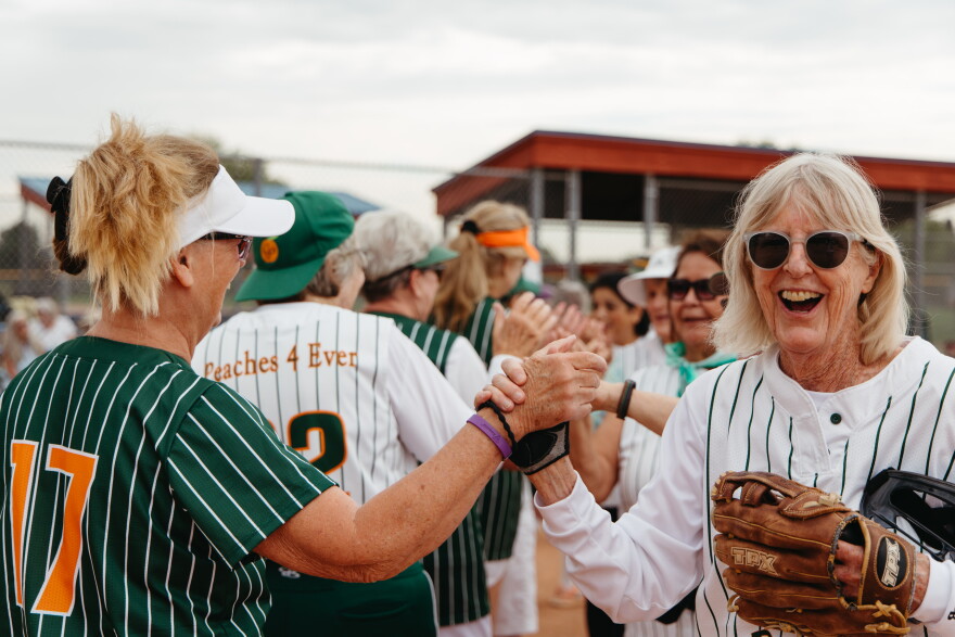 One senior woman in a green baseball jersey and a white visor high fives the hand of another senior woman in a white jersey holding a baseball glove in her other hand. She is smiling. The women are both in lines walking next to each other, and behind them are other women giving out high fives in the line.