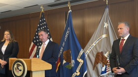 Federal officials stand in front of flags at a podium at a news conference. The podium has the Justice department's logo on it. 