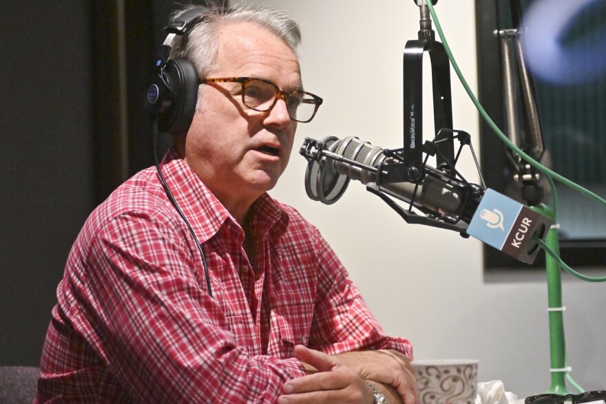 A man sitting in a radio studio sits behind a microphone and is talking.