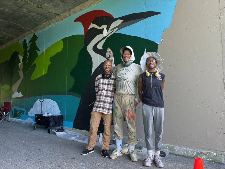 The three artists pose in front of a tunnel wall with their mural, which shows a woodpecker, a hill and a pond. A wagon, camping chair and construction cone are also visible. 