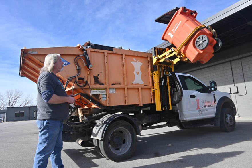 A man stands outside on a paved surface outside a warehouse. He is standing next to a truck that has is lifting a large orange trash can to empty into the back of the truck.