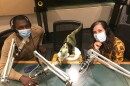 A photo of two people at microphones, wearing masks, and a stuffed dinosaur in the middle