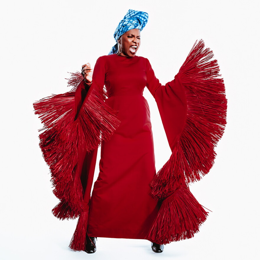 A woman in a dramatic, tassled. floor-length red dress and blue head turban, singing forcefully