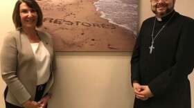 Dr. Deborah Beidel and Father José Rodríguez stand in front of a picture of the word "Restores" written on a beach in Puerto Rico. Photo: Danielle Prieur