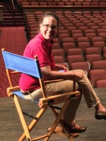 A woman wearing glasses  in a red polo shirt looks over her shoulder while sitting in a blue directors chair on a stage in an empty theater.