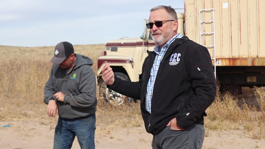 Yuma county commissioner Scott Weaver gestures with a hand while speaking in the foreground. Nathan Weathers checks his watch behind him. A truck is parked in a plain behind them.