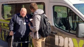 Two men stand facing each other, one older with a walker, in front of a vehicle with the words "DAV" written on its side.