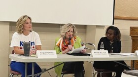 Three women sit at a table with name placards, one holding a microphone.