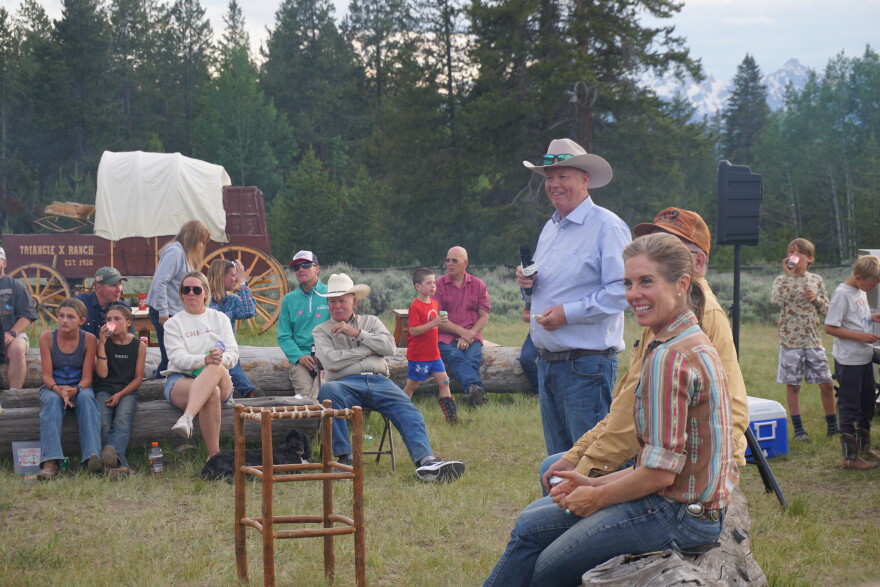 A woman wearing jeans and a western shirt smiles, as others sit on logs outside to listen to stories.