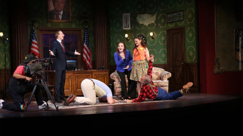 Six people appear on a set made to look like a governor's office. Two men are on the floor, two women stand, one man stands with his hands stretched out to either side of him, and another man kneels behind a camera. 