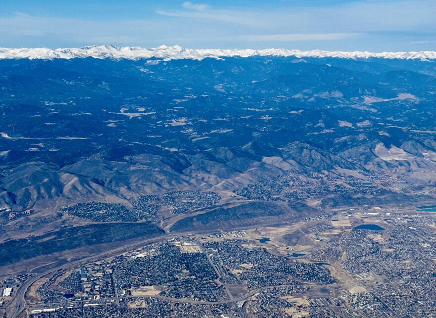 Dry mountains and hills are pictured next to sprawling cities as seen from an aerial photograph. A small ribbon of snow capped mountains are in the background. 