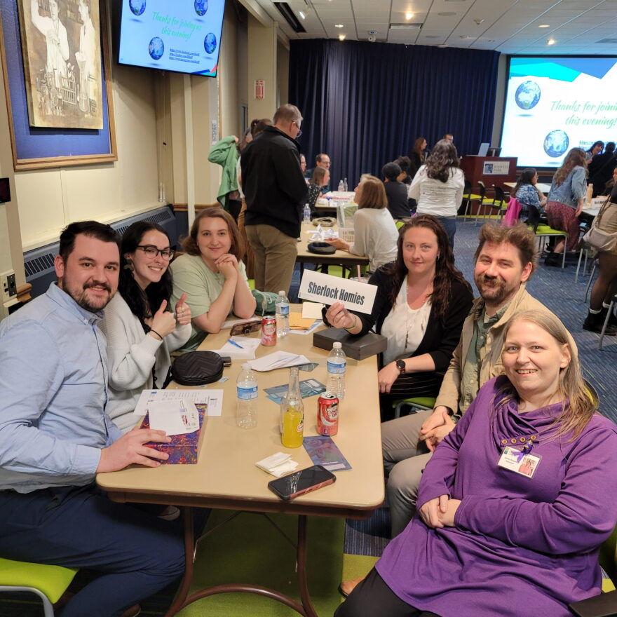 A group of people sit at a table and smile. One person holds up a sign of their team name that reads "Sherlock Homies".
