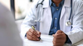 A doctor, with a stethoscope around his neck, writes on a paper on a desk